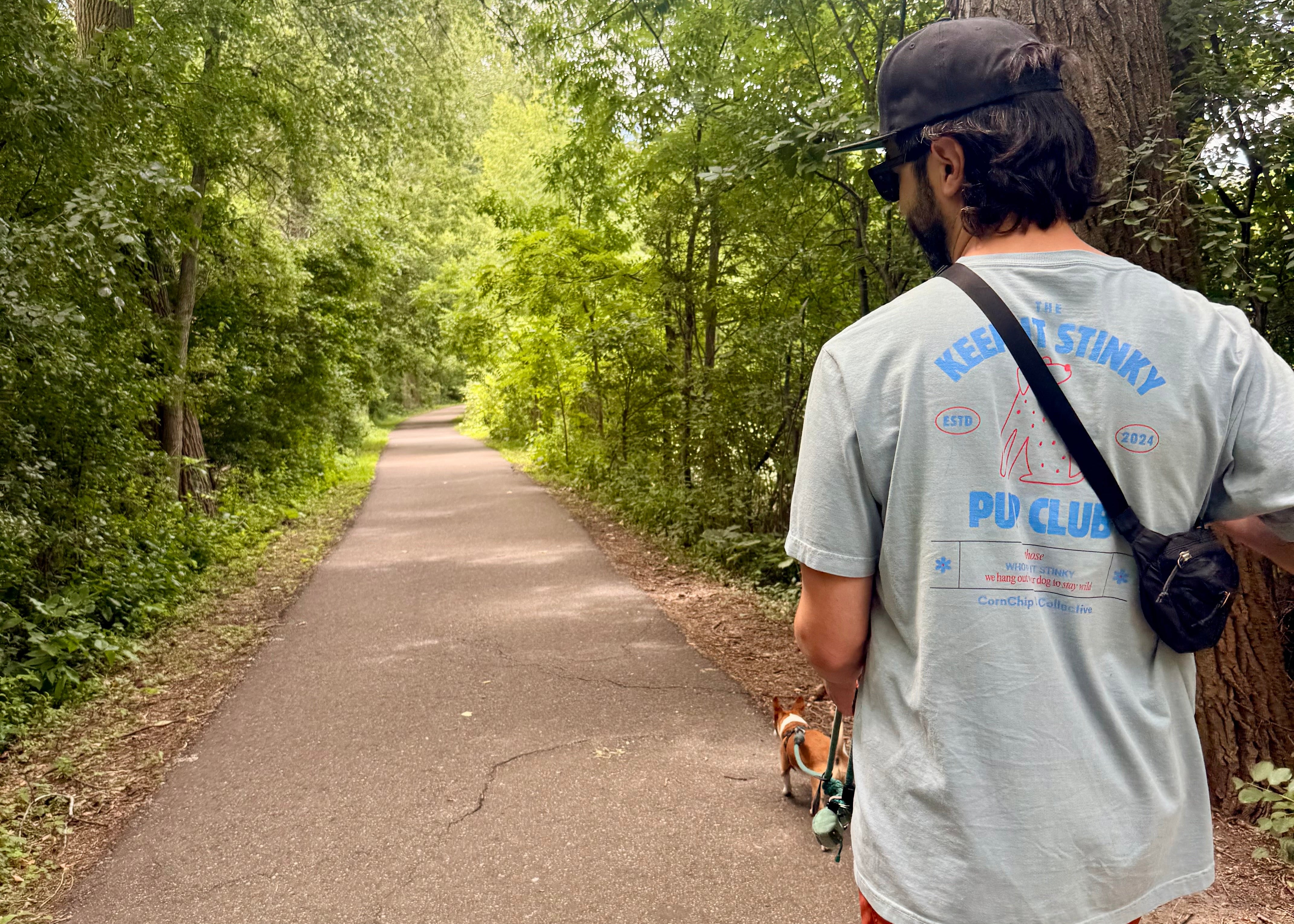 A man wearing a light blue t-shirt with blue 'KEEP IT STINKY PUP CLUB' text, orange pants, and a black cap walks along a paved forest path with a small dog. 