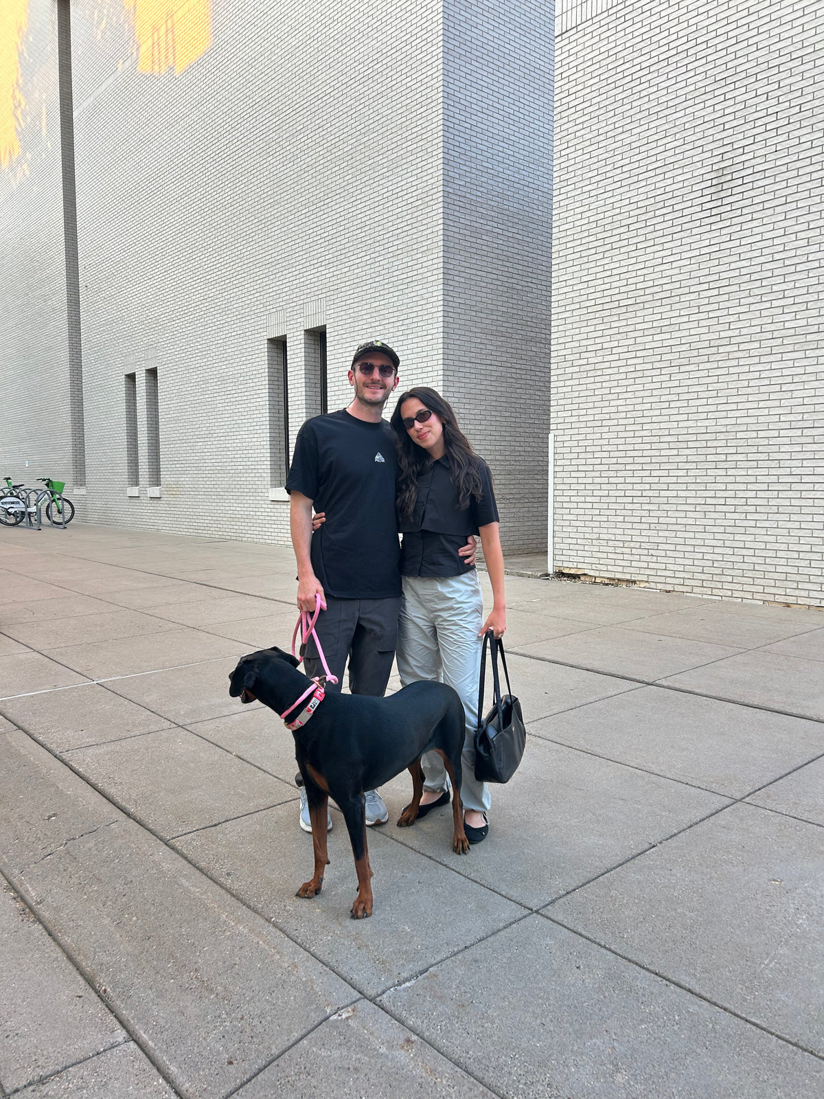 A man and a woman standing outdoors on a paved walkway next to a large gray brick building, with a black dog wearing a pink harness beside them. The man wears a black t-shirt and dark pants, and the woman wears a black top and light pants.